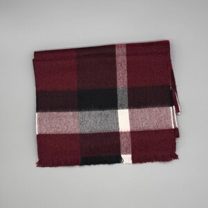 May include: A rectangular scarf featuring a plaid pattern in shades of deep burgundy, black, white, and gray. The fabric has a soft, woven texture with a fringed edge along the bottom, displaying a classic grid design with intersecting lines.