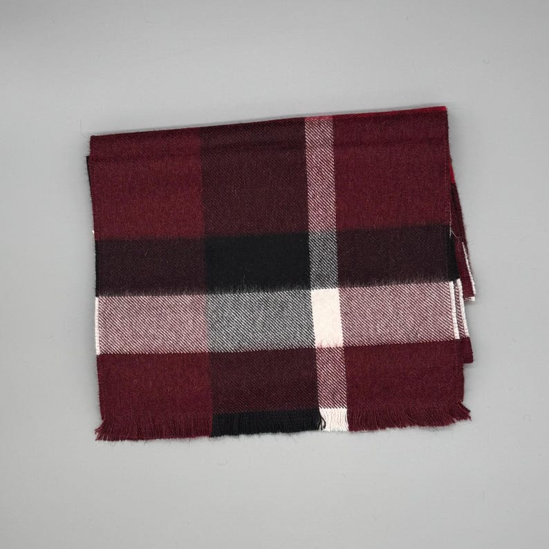May include: A rectangular scarf featuring a plaid pattern in shades of deep burgundy, black, white, and gray. The fabric has a soft, woven texture with a fringed edge along the bottom, displaying a classic grid design with intersecting lines.