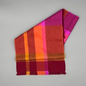 May include: A rectangular scarf featuring a vibrant plaid pattern in shades of magenta, bright orange, deep red, and yellow. The fabric displays a woven texture with a fringed edge at the bottom, showcasing a bold color-blocked design throughout.