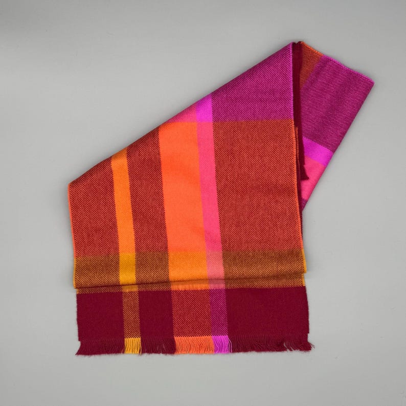 May include: A rectangular scarf featuring a vibrant plaid pattern in shades of magenta, bright orange, deep red, and yellow. The fabric displays a woven texture with a fringed edge at the bottom, showcasing a bold color-blocked design throughout.