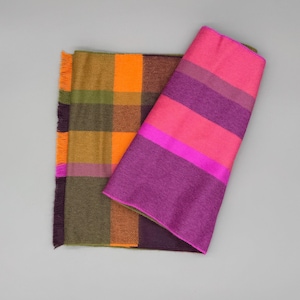 May include: A reversible scarf featuring two distinct patterns. One side displays a large plaid design in shades of orange, olive green, and dark purple. The other side shows bold horizontal stripes in vibrant hot pink and deep purple tones.