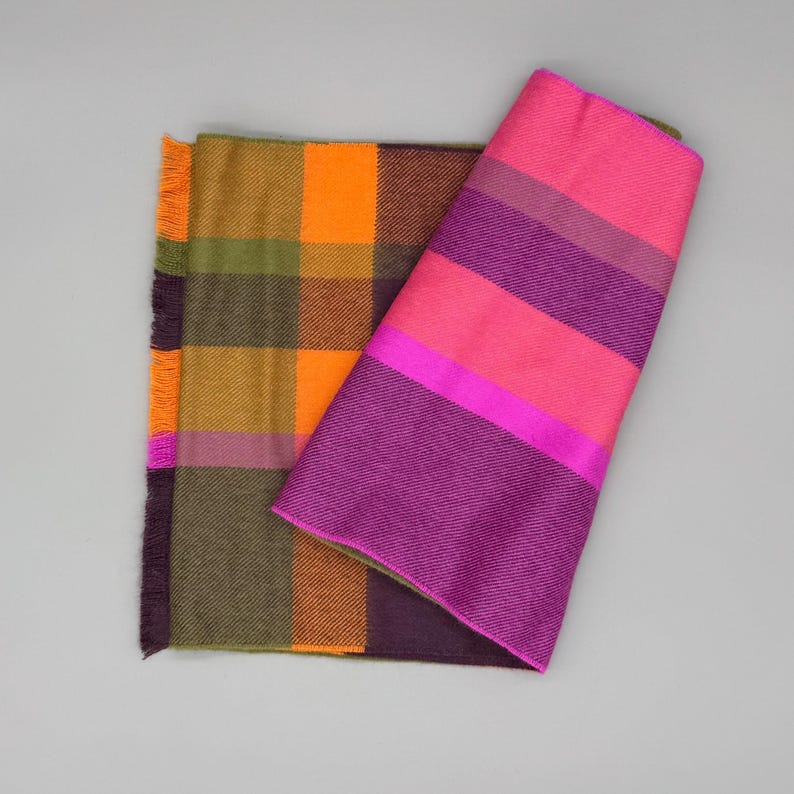 May include: A reversible scarf featuring two distinct patterns. One side displays a large plaid design in shades of orange, olive green, and dark purple. The other side shows bold horizontal stripes in vibrant hot pink and deep purple tones.