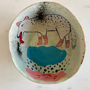Può includere: Una ciotola in ceramica con un design dipinto a mano di un animale bianco macchiato che indossa calzini rosa in piedi su una nuvola blu. La ciotola ha una nuvola rosa e bianca con punti neri e uno sfondo bianco.