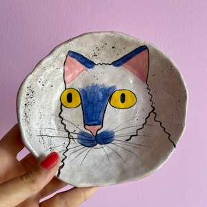 Può includere: Un piatto in ceramica fatto a mano con un design a forma di volto di gatto stravagante. Il volto del gatto è dipinto con occhi gialli, naso e orecchie blu e orecchie rosa. Il piatto ha uno sfondo bianco sporco macchiettato.