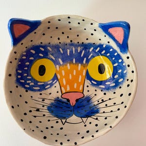 Può includere: Una ciotola in ceramica bianca con un viso di gatto blu e giallo dipinto su di essa. Il gatto ha orecchie rosa, un naso nero e baffi bianchi. La ciotola è ricoperta di pois neri.
