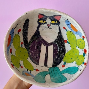Può includere: Ciotola in ceramica dipinta a mano con un gatto bianco e nero con occhi gialli, circondato da cactus verdi e uccelli blu. La ciotola ha uno sfondo bianco macchiettato. Un design artistico unico. Un pezzo decorativo e stravagante.