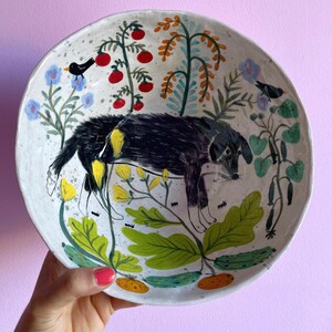 Op de afbeelding: Handbeschilderde keramische plaat met een ontwerp van een zwarte hond, rode bessen, blauwe bloemen en groene bladeren. De plaat heeft een gespikkelde, gebroken witte achtergrond. De plaat wordt vastgehouden door een persoon met een roze gelakte nagel.