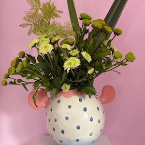 Könnte beinhalten: Eine runde Keramikvase mit weißem Grund, blauen Punkten und einem hellblauen Farbverlauf am Boden. Sie hat zwei rosa, ohrförmige Seitengriffe und einen gewellten rosa Rand. Die Vase ist mit einem Strauß aus grünen Chrysanthemen und Farnen gefüllt.