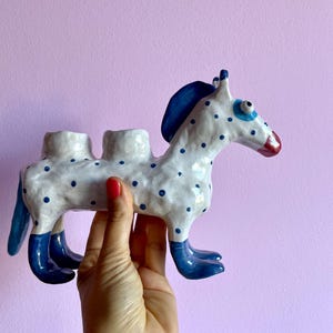 May include: A whimsical ceramic horse figurine, white with blue polka dots, a blue mane, and blue boots. The horse has a red nose and is designed to hold two items. The figurine is held against a light purple background.