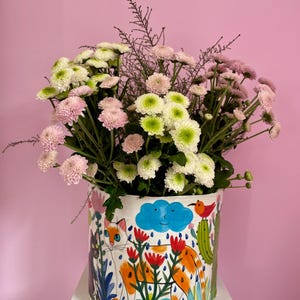 Può includere: Un vaso cilindrico in ceramica con un disegno dipinto a mano raffigurante un gatto, fiori e una nuvola. Contiene un bouquet di crisantemi rosa e bianchi. Il vaso è su una superficie bianca contro uno sfondo rosa.