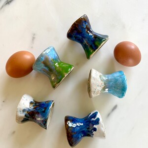 Può includere: Sei porta uova in ceramica in varie tonalità di blu, verde e marrone con macchie bianche. I porta uova sono in diversi design e sono disposti su una superficie di marmo bianco.