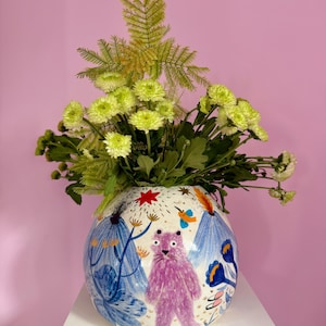 Könnte beinhalten: Eine runde weiße Keramikvase mit einer handgemalten Illustration eines violetten Bären, blauen Bergen und orangefarbenen Akzenten. Die Vase ist mit einem Strauß aus hellgrünen Chrysanthemen und federartigem grünem Blattwerk gefüllt.