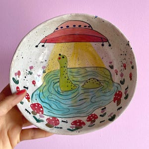 Peut inclure: Une assiette en céramique fantaisiste représentant une scène de lac avec une créature verte, possiblement le monstre du Loch Ness, sous un OVNI rouge. L'assiette est décorée de champignons rouges et blancs et de petites fleurs, sur fond blanc moucheté.