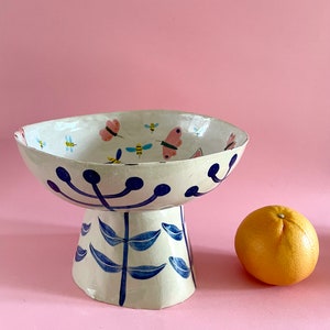 Può includere: Una ciotola in ceramica bianca con un motivo floreale blu e bianco e farfalle e api dipinte su di essa. La ciotola è su un piedistallo con un motivo floreale blu e bianco.