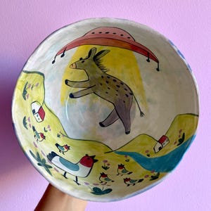 May include: A hand-painted ceramic bowl with a quirky design. It depicts a donkey leaping under a red UFO, set against a yellow sun. The bowl's rim shows a landscape with birds, houses, and a blue river.