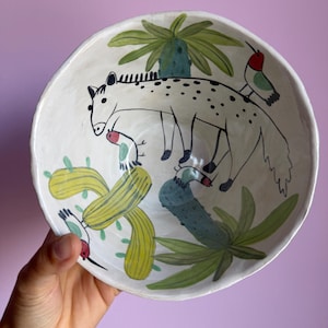 May include: A white ceramic bowl featuring hand-painted illustrations of a spotted animal, green cacti, palm-like plants, and small birds with red heads. The bowl has an irregular, organic rim and a smooth, glossy finish throughout the interior.