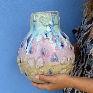May include: A ceramic vase with a bulbous shape featuring a light green top, a pink middle section, and a speckled beige base. The surface is decorated with raised relief designs of clouds, stars, and raindrops in blue and white glaze drips.