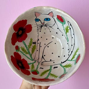 Può includere: Scodella in ceramica bianca con un design dipinto a mano di un gatto bianco con macchie nere seduto in un campo di papaveri rossi. Il gatto ha gli occhi azzurri e un naso nero.