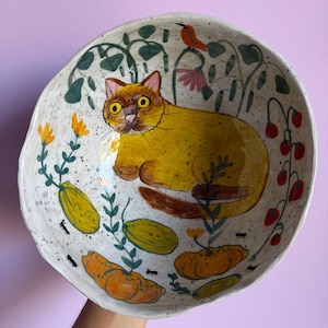 May include: A hand-painted ceramic bowl with an irregular, organic rim. The interior features a large yellow cat surrounded by orange pumpkins, green vines, small red berries, and a tiny orange bird, all set against a speckled off-white background.
