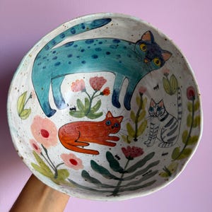 Op de afbeelding: Een ronde keramische kom met een gespikkeld gebroken wit glazuur, voorzien van handgeschilderde illustraties van drie katten in blauw, oranje en grijs. Het ontwerp bevat kleurrijke bloemmotieven, groen gebladerte en kleine insecten.