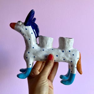 Peut inclure: Figurine de cheval en céramique fantaisiste, corps blanc à pois bleus, détails bleus sur les pattes et la crinière, et queue orange. Deux ouvertures sur le dos. La figurine est tenue par une personne.