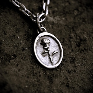 May include: Silver pendant necklace featuring an oval pendant with a raised rose design. The pendant is attached to a silver chain. The necklace is displayed on a dark, textured surface, highlighting the jewelry's details.