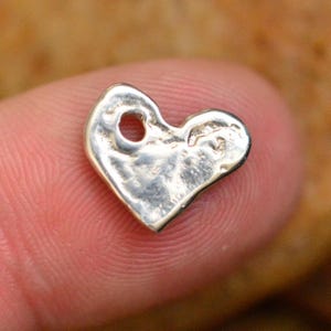 Può includere: Un piccolo ciondolo a forma di cuore color argento con un foro nella parte superiore. Il ciondolo ha una superficie strutturata ed è probabilmente in metallo. Il cuore è tenuto tra due dita, suggerendo che si tratti di un ciondolo o di un piccolo elemento decorativo.