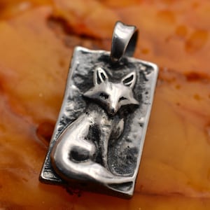 Könnte beinhalten: Silberfarbener Anhänger mit einem detaillierten Fuchsmotiv. Der Fuchs sitzt in einem rechteckigen Rahmen. Der Anhänger hat eine Schlaufe zum Aufhängen an einer Kette und eine strukturierte, antike Oberfläche.
