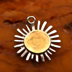 Op de afbeelding: Een zilverkleurige zonnehanger met een goudkleurig midden. De hanger heeft een lus aan de bovenkant om aan een ketting of koord te bevestigen. De zonnestralen zijn gelijkmatig verdeeld rond de centrale schijf.