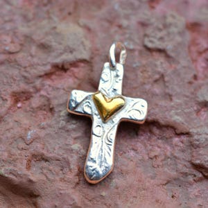 Può includere: Ciondolo in argento a forma di croce con un cuore dorato al centro. La croce presenta un design ornato e inciso e un anello nella parte superiore per una catena. Il ciondolo è esposto su una superficie strutturata marrone-rossastra.