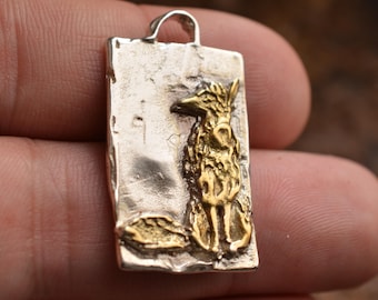 Könnte beinhalten: Rechteckiger Silberanhänger mit einem goldenen Fuchsmotiv. Der Fuchs sitzt, mit detailliertem Fell und spitzer Schnauze. Der Anhänger hat oben eine Schlaufe für eine Kette. Der Hintergrund ist strukturiert.