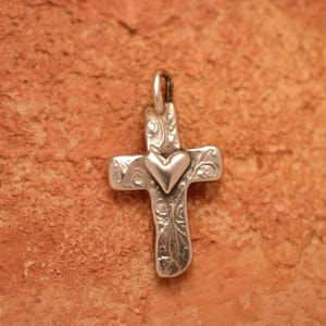 Può includere: Ciondolo color argento a forma di croce con un cuore al centro. La croce presenta un design inciso e ornato. Il ciondolo è esposto su uno sfondo strutturato marrone-rossastro.