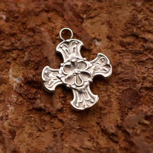 Puede incluir: Colgante de cruz plateada con un diseño floral en el centro y detalles ornamentales en cada brazo. Tiene un pequeño lazo en la parte superior para colgarlo. El colgante se encuentra sobre un fondo marrón texturizado.