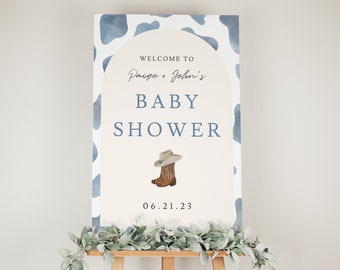 May include: A baby shower sign on an easel. The sign has a light beige background with a blue cow print border. The text reads "Welcome to Paige + John's Baby Shower" with a cowboy boot and hat illustration. The date is 06.21.23.