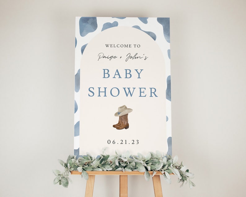 May include: A baby shower sign on an easel. The sign has a light beige background with a blue cow print border. The text reads "Welcome to Paige + John's Baby Shower" with a cowboy boot and hat illustration. The date is 06.21.23.