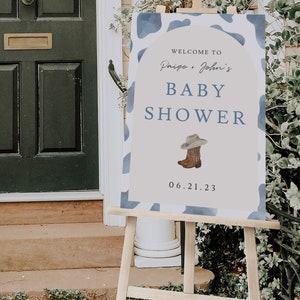 May include: A baby shower sign on a wooden easel. The sign has a blue and white cow print border and reads "Welcome to Paige + John's BABY SHOWER" with a cowboy hat and boots graphic. The date is June 21, 2023.