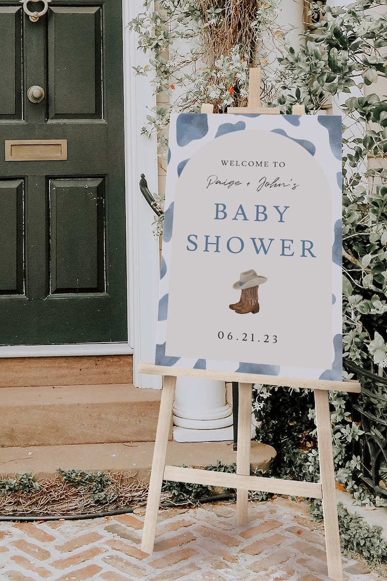 May include: A baby shower sign on a wooden easel. The sign has a blue and white cow print border and reads "Welcome to Paige + John's BABY SHOWER" with a cowboy hat and boots graphic. The date is June 21, 2023.