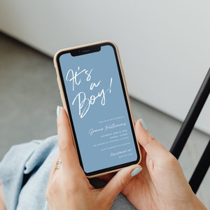 May include: A smartphone displaying a baby shower invitation with a blue background and white text that reads "It's a Boy!" The invitation includes the name "Grace Williams", the date "Saturday, April 8, 2023", the time "10:00am-12:00pm", the location "148 Golden Dr Pasadena, CA 91104", the RSVP information "RSVP to Brenda 628-129-8760", and the website "Registered at Babylist.com".