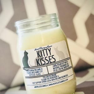 May include: A glass jar candle with a white label that says "Novel Design Home Kitty Kisses". The label also says "Made in New Orleans & poured into hand-cut recycled glass" and "100% Vegan Soy Wax & Natural Fragrance Oil 20 oz. | 190 hr. Burn Time". The label also includes the text "Follow us online to join the movement & help support other local New Orleans businesses @noveldesignnola | www.noveldesignnola.com".