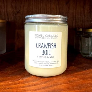 May include: A white candle with a blue label that reads 'Novel Design Home Crawfish Boil' and includes details about the candle such as being made with 100% vegan soy wax and having a natural fragrance. The candle is 20 oz and has a 190+ hour burn time. The label also encourages customers to support local New Orleans businesses.