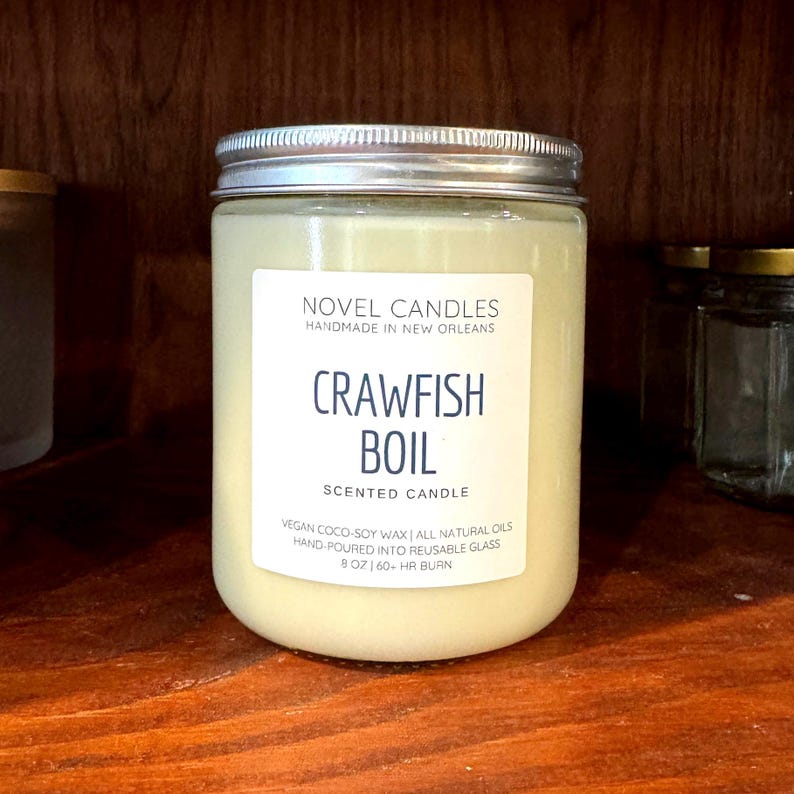 May include: A white candle with a blue label that reads 'Novel Design Home Crawfish Boil' and includes details about the candle such as being made with 100% vegan soy wax and having a natural fragrance. The candle is 20 oz and has a 190+ hour burn time. The label also encourages customers to support local New Orleans businesses.