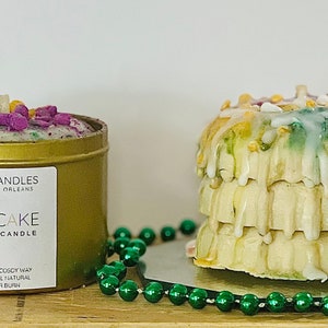 May include: A gold candle tin with a green, purple, and gold king cake design. The text on the tin reads "Novel Candles Made in New Orleans King Cake Scented Candle 100% Vegan Coconut Wax Handmade | All Natural 4 oz (113g) Burn". A king cake candle is next to the tin, with green, purple, and gold icing and sprinkles.