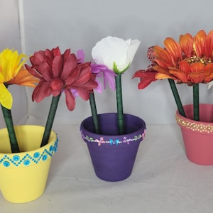 May include: Three flower pots with colorful flowers and decorative trim. The pots are painted yellow, purple, and pink. The flowers are red, orange, white, and yellow.