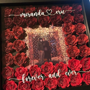 May include: A framed shadow box featuring a photograph surrounded by red paper roses. The names "miranda" and "eric" are written above the photo, with "forever and ever" below. The frame is black.