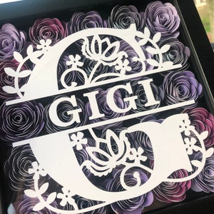 Op de afbeelding: Een ingelijst bloemenkunstwerk met papieren rozen in tinten paars en wit. Het ontwerp bevat de naam "GIGI" in een decoratief wit lettertype, met bloemversieringen. De lijst is zwart.