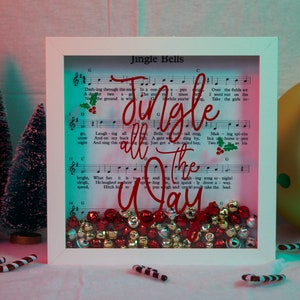 Puede incluir: Decoración navideña enmarcada con partituras y la frase "Jingle all the Way" en rojo. El marco contiene campanas rojas y doradas, rodeado de pequeños árboles de Navidad y bastones de caramelo. El fondo tiene un degradado rosa y turquesa.