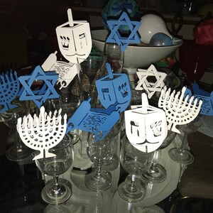 May include: A set of blue and white paper cutouts of menorahs, dreidels, and Stars of David, attached to wine glasses. The cutouts are a festive decoration for a Hanukkah celebration.