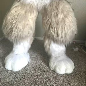 May include: Furry costume legs in light brown and white, featuring large, white paws. The legs are covered in long, shaggy fur, changing from light brown to white at the feet. The paws are large and rounded.