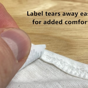 May include: A white t-shirt with a label that tears away easily for added comfort. The label is partially torn off, revealing the white fabric underneath.
