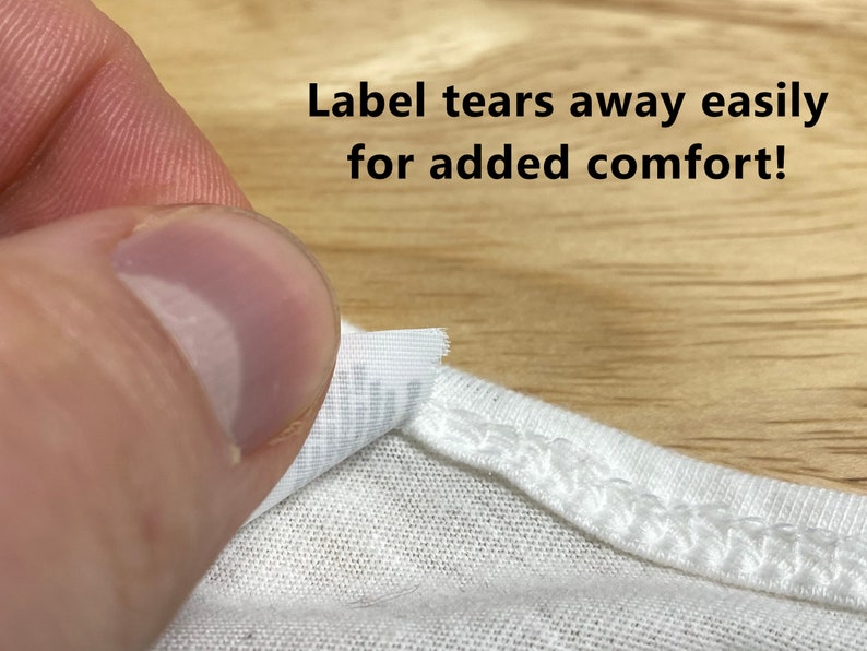 May include: A white t-shirt with a label that tears away easily for added comfort. The label is partially torn off, revealing the white fabric underneath.